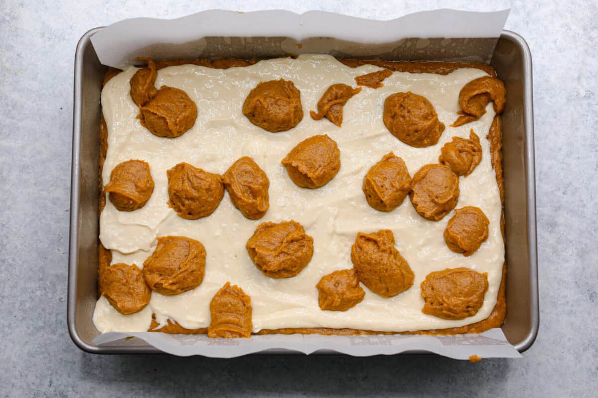 Overhead shot of baking pan with layer of pumpkin roll mixture, topped with cream cheese mixture with dollops of more pumpkin roll mixture on top. 