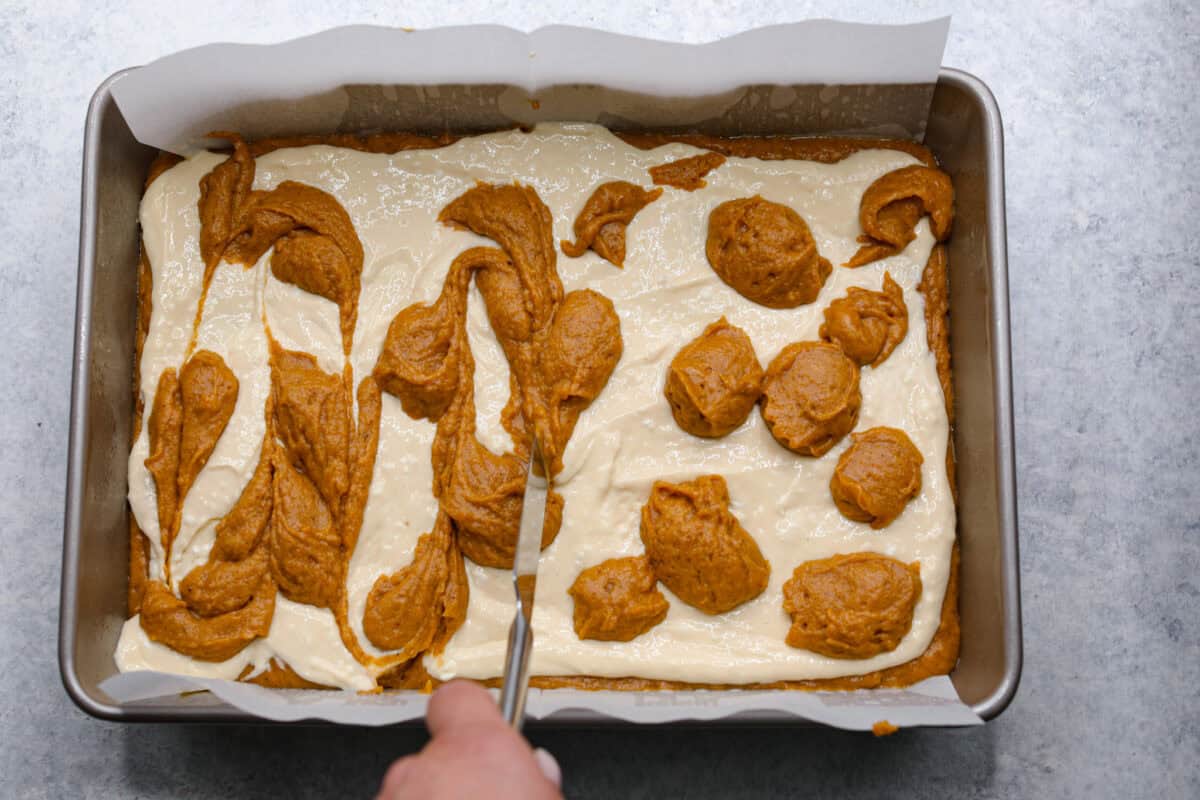 Overhead shot of someone swirling the dollops of pumpkin roll into the cream cheese layer. 