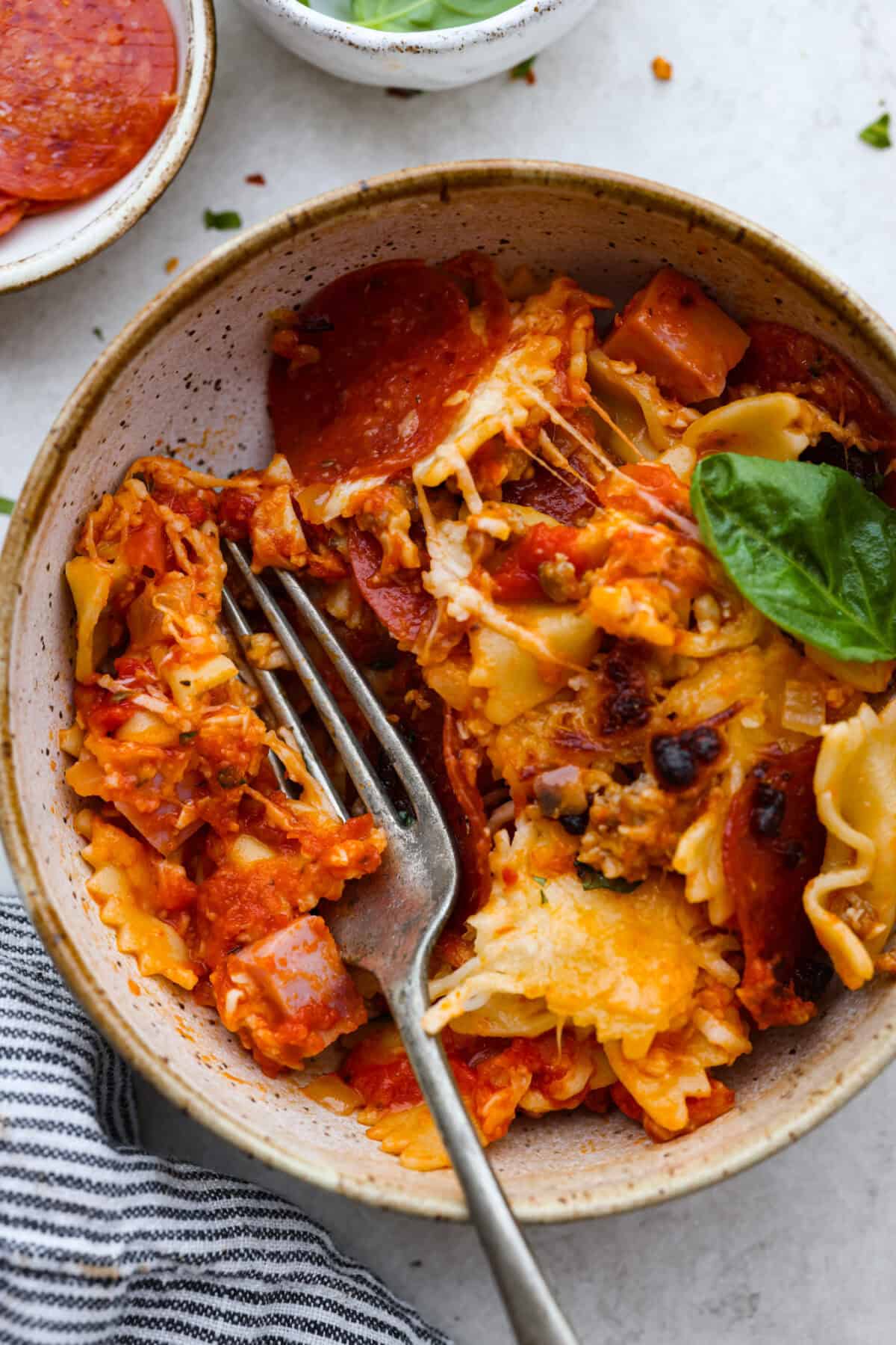 Overhead shot of a bowl with a serving of cheesy pizza casserole in it with a fork and basil leaf. 