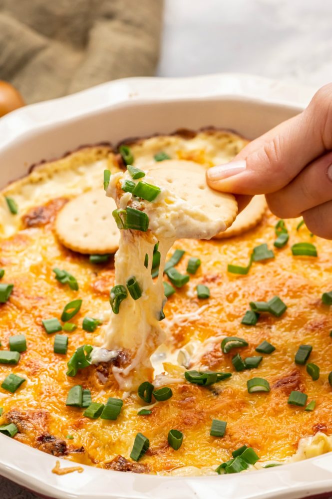 Asiago dip in a white dish with ritz crackers being dipped in the asiago. 