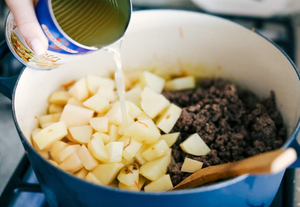 Potatoes, ground beef and vegetable in a pot with chicken broth being poured over it. 