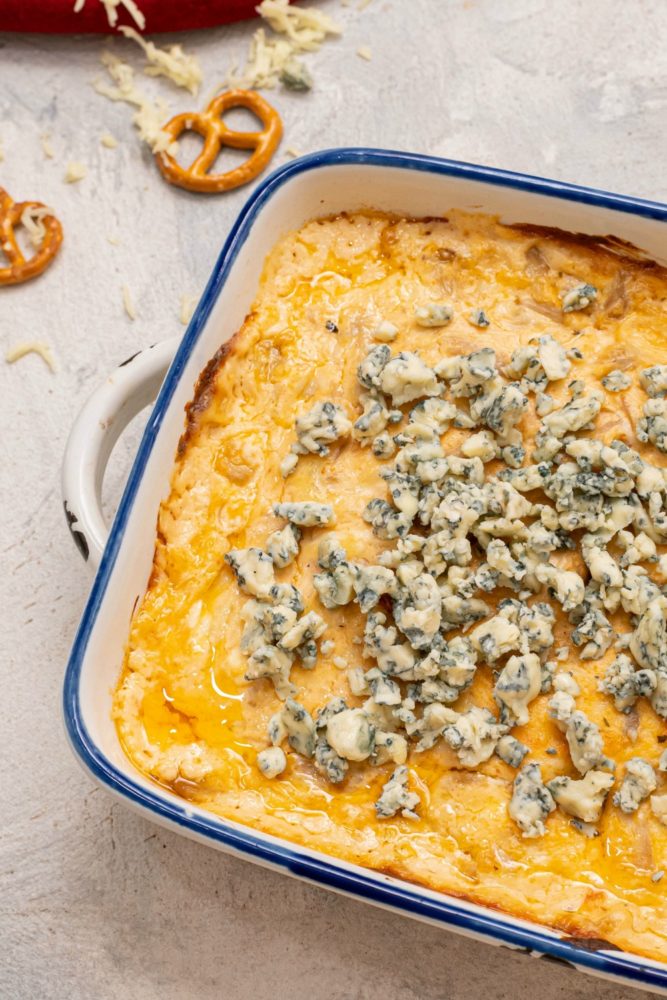 A blue and white dish with buffalo dip with blue cheese crumbles on top. Two pretzels lay on the counter beside the pan.