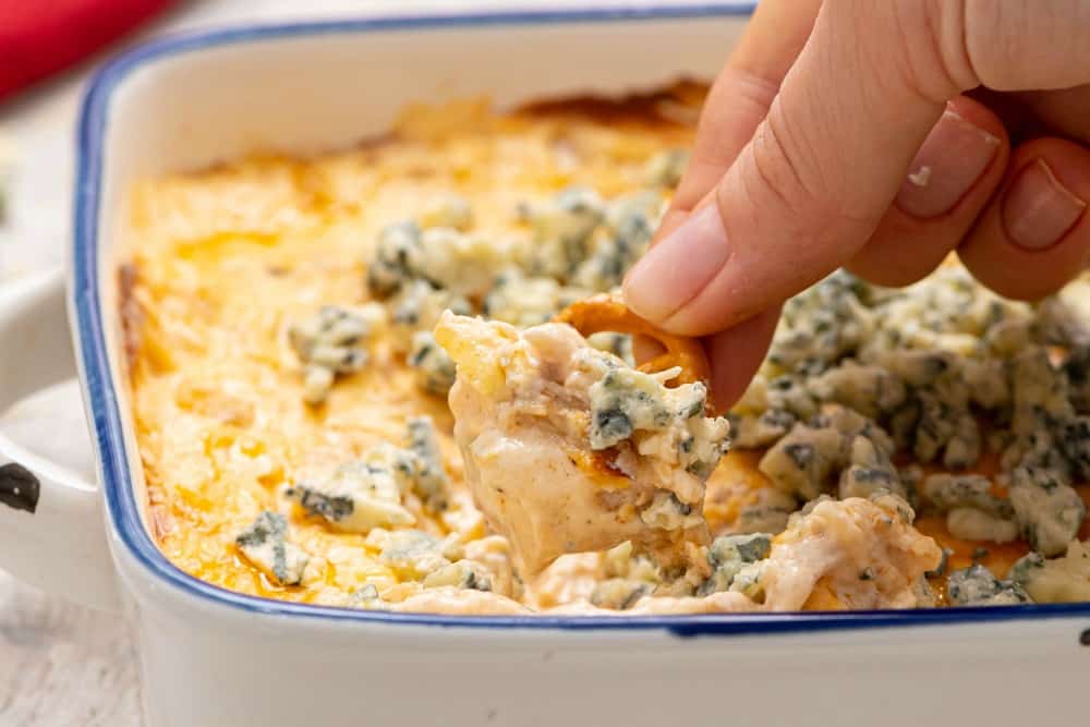 A hand with a chip in it is dipping some of the buffalo dip out of a blue and white dish.