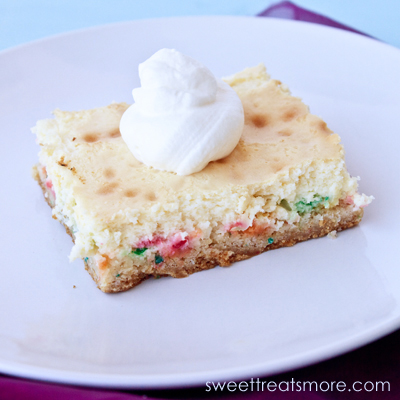 On funfetti cheesecake square with a dollup of whipped cream on top, sitting on a round white plate.