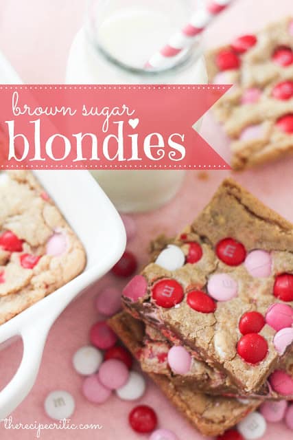 A stack of brown sugar blondies with pink, white, and red m and m's sit beside a glass of milk with a red and white striped straw and another brownie and a white pan of brownies.