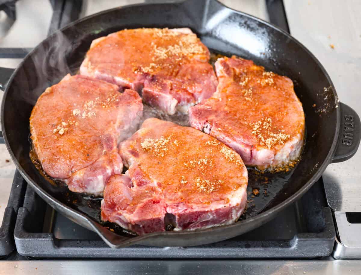 Overhead shot of pork chops searing in a skillet on a stove top. 