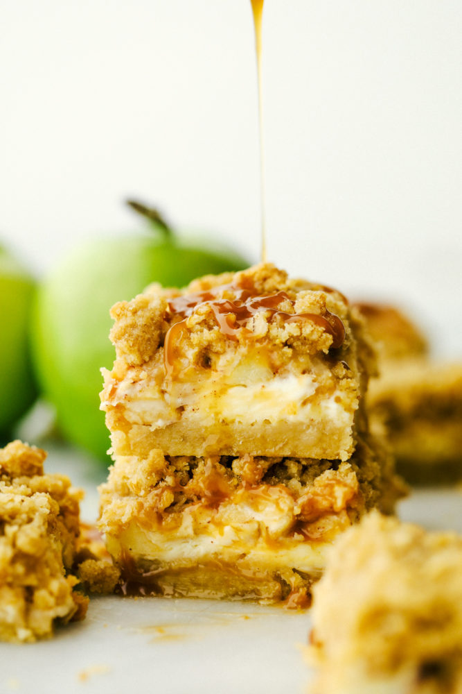 Two caramel apple cheesecake bars stacked on top of each other with caramel being drizzled over top.