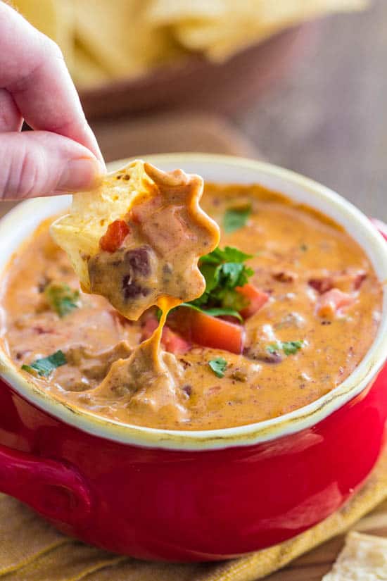 Close up of Chili Queso in a red serving cup with a person dipping a tortilla chip into it. 