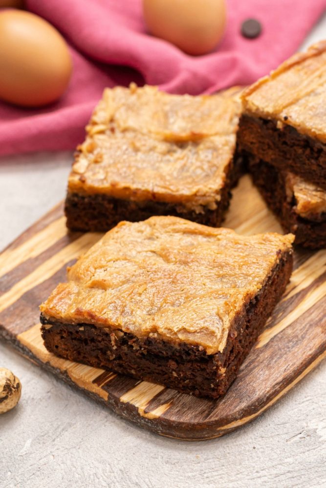 Peanut Butter Cheesecake Brownie Bars on a wooden cutting board with two eggs in the background. 