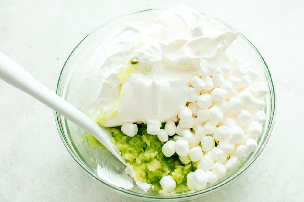 Pistachio pudding salad ingredients in a glass bowl being stirred with a white spatula. 