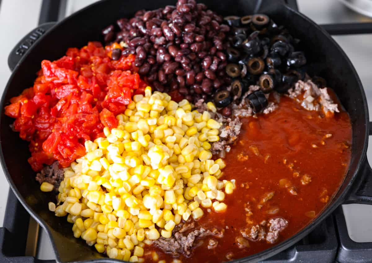 Overhead shot of cooked beef and onions with enchilada sauce, corn, olives, tomatoes and beans on top. 