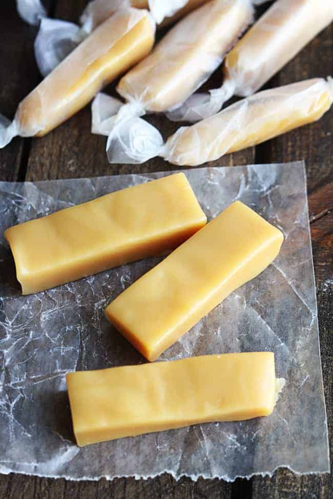 Unwrapped microwavable Caramels with three of them on a parchment paper with wrapped microwavable Caramels behind them in the photo.