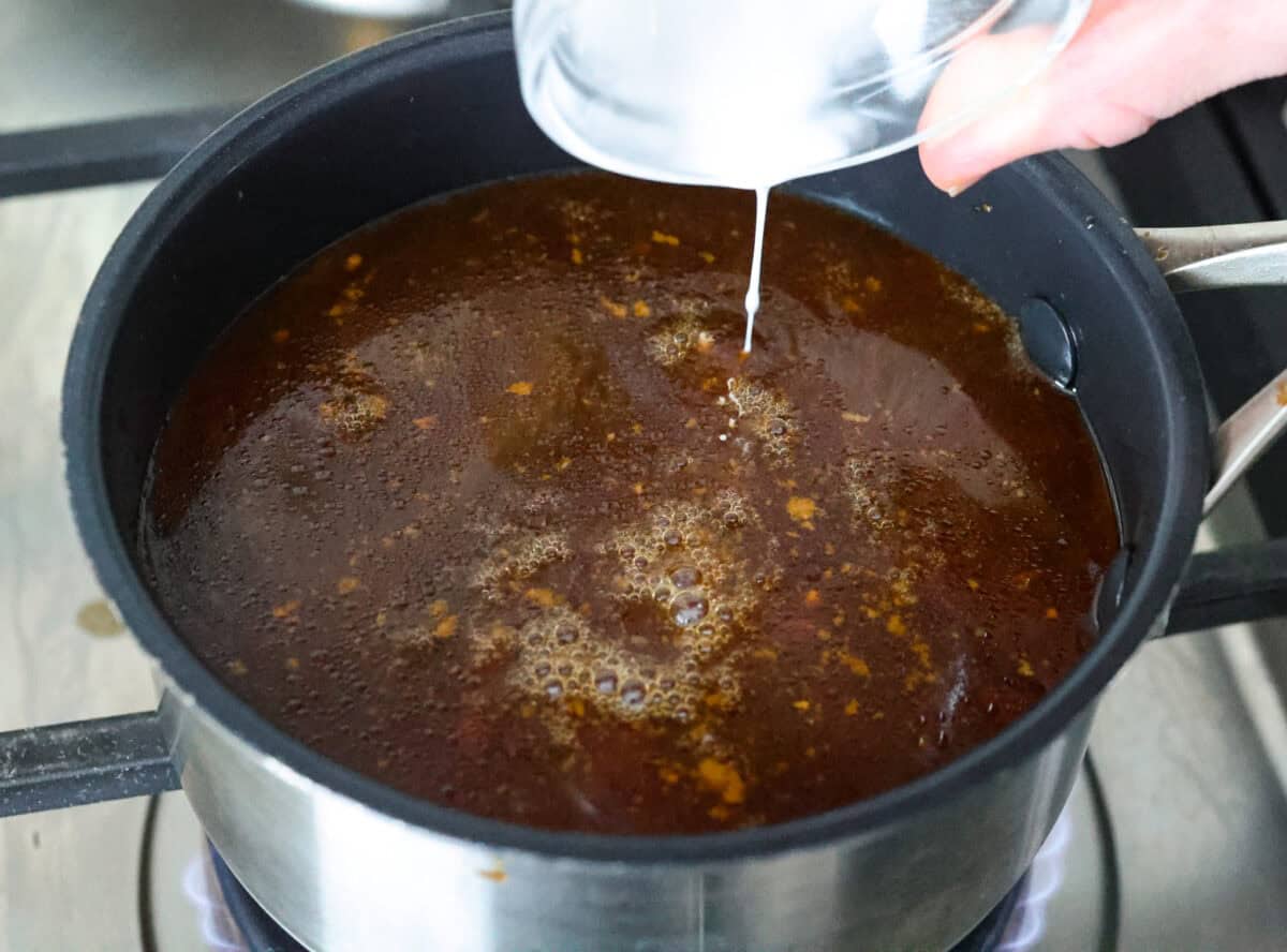 Angle shot of someone pouring cornstarch mixture into the sauce in a pot on the stove. 