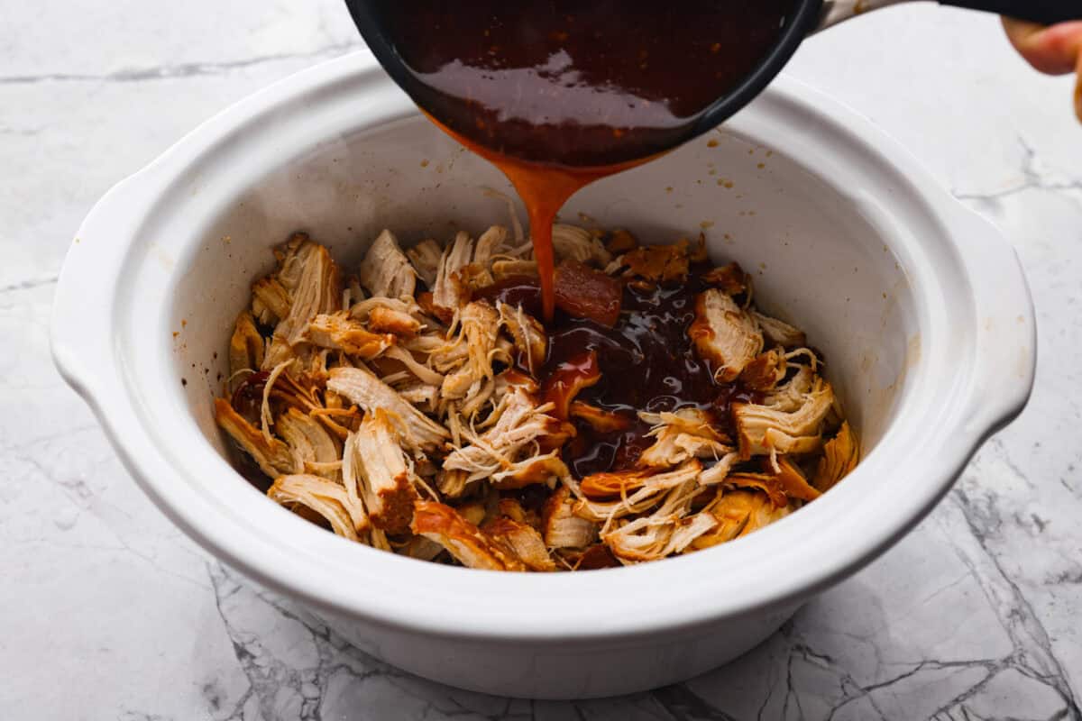 Angle shot of someone pouring honey teriyaki sauce over the shredded chicken in the crockpot. 