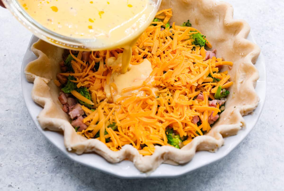 Overhead shot of someone pouring the egg mixture into a pie crust with the ham, broccoli and shallot mixture topped with cheese. 