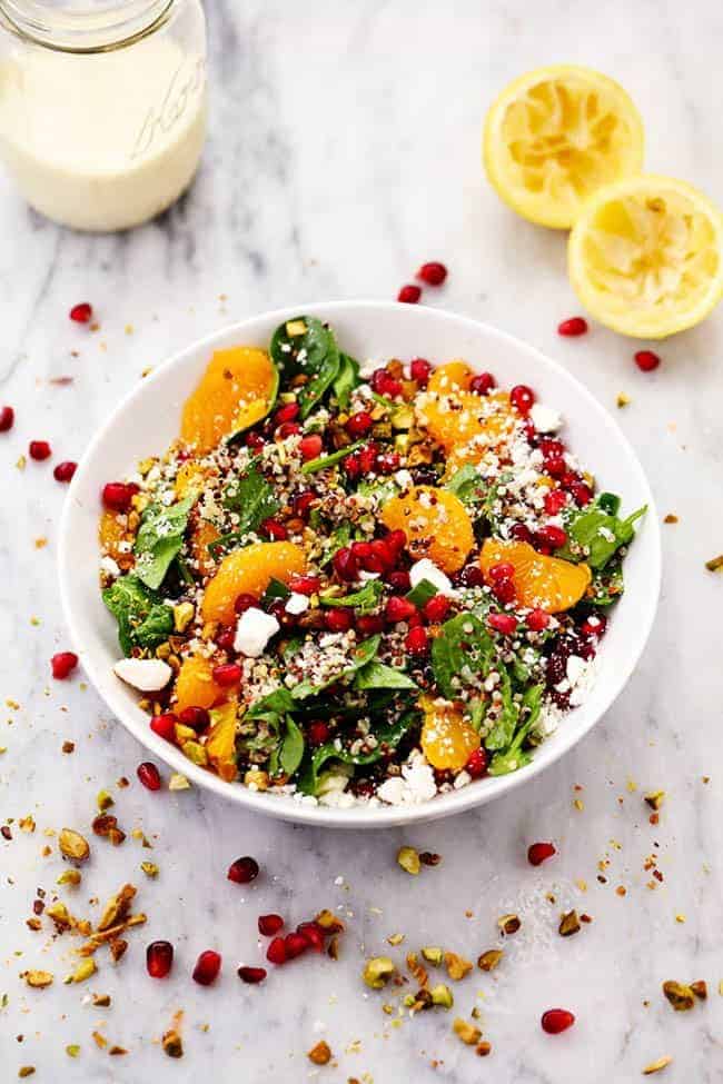 WINTER POMEGRANATE ORANGE QUINOA SALAD WITH CREAMY LEMON GREEK YOGURT DRESSING IN A WHITE BOWL WITH TWO FRESHLY SQUEEZED LEMONS ON THE SIDE.  