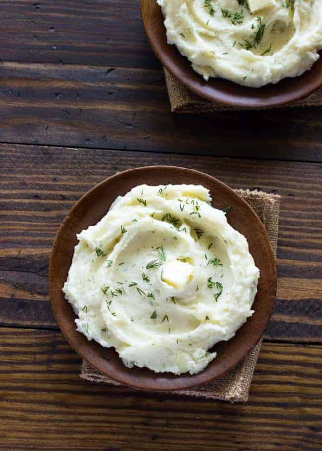 Overhead photo of Slow Cooker Ranch Mashed Potatoes in a brown bowl with a brown cloth under, placed on a wooded table. 