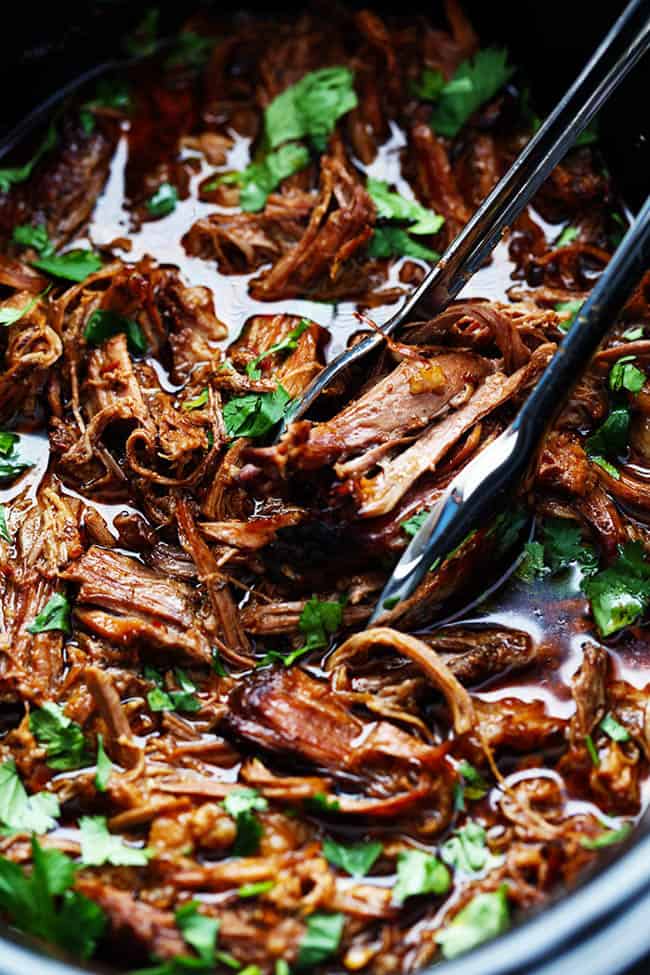 Beef barbacoa in a slow cooker with tongs pulling up a bit of the beef to be shown in the photo.