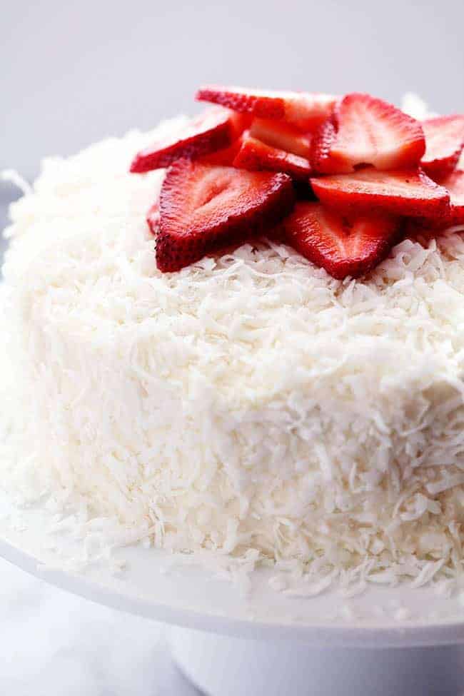 Strawberry Coconut Cream Cake with Coconut Cream Cheese Frosting and fresh cut strawberries on top displayed on a white cake stand. 