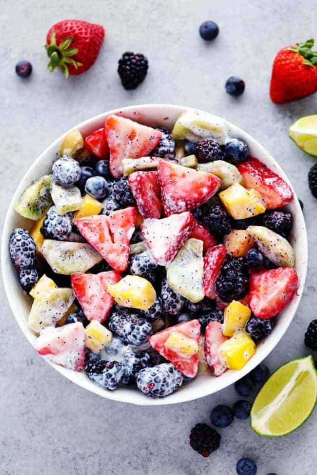 Creamy Poppyseed Fruit Salad in a white bowl with fresh lemon, blueberries, blackberries and strawberries on the side. 