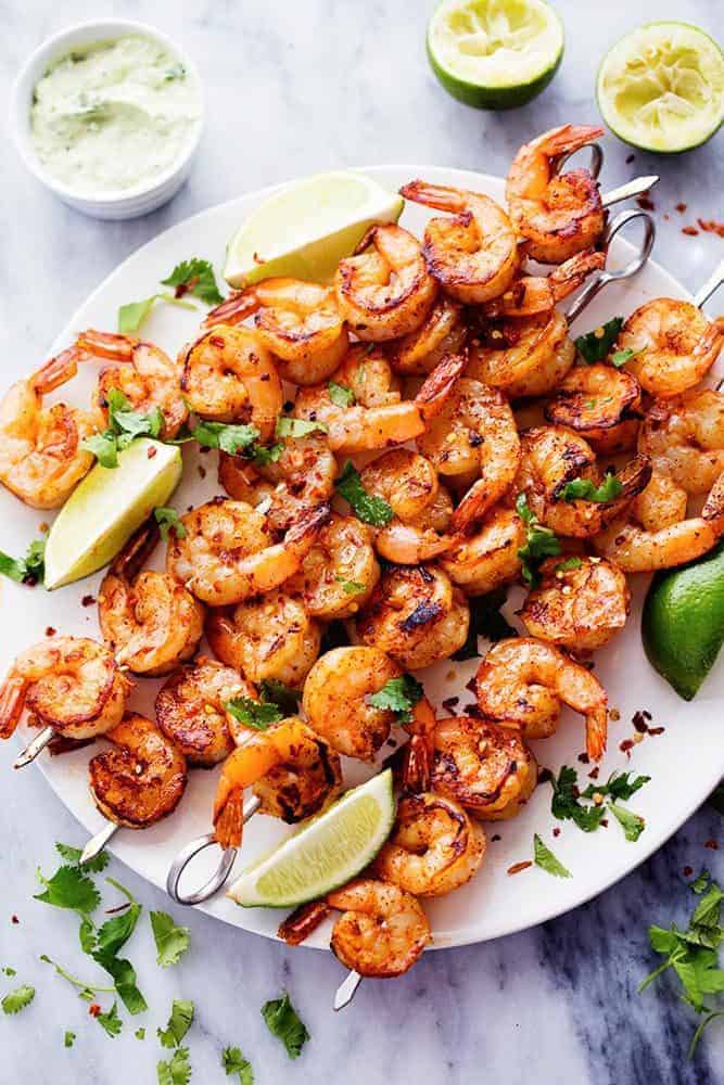 Grilled Spicy Lime Shrimp with Creamy Avocado Cilantro Sauce on skewers on a white plate with fresh limes on the side. 