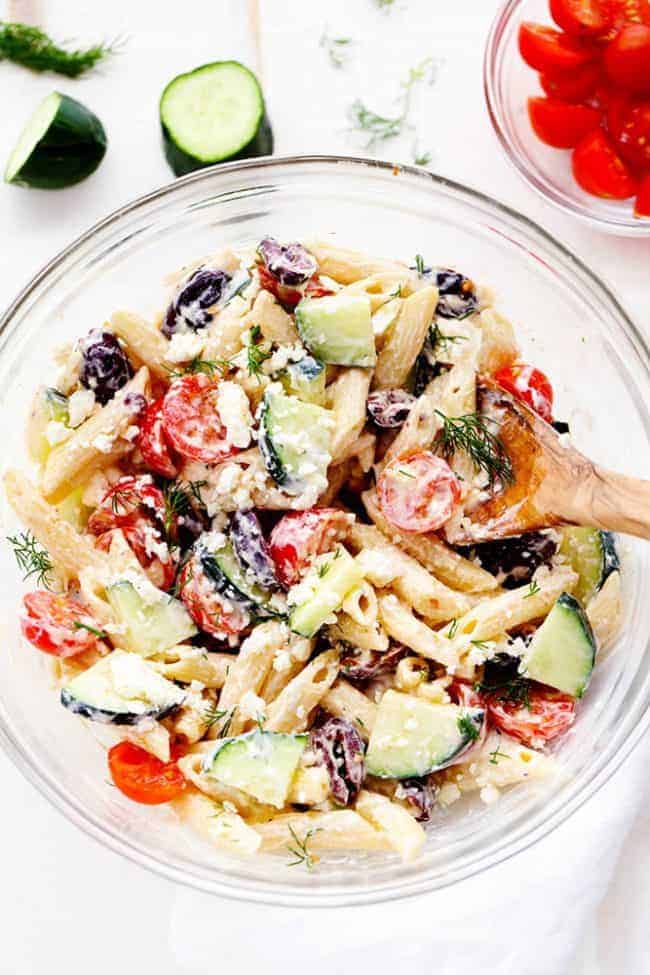 Overhead view of Greek Tzatziki Pasta Salad in a clear mixing bowl with a wooden spoon. There is a small clear bowl of tomatoes in the corner as well as a sliced cucumber. 