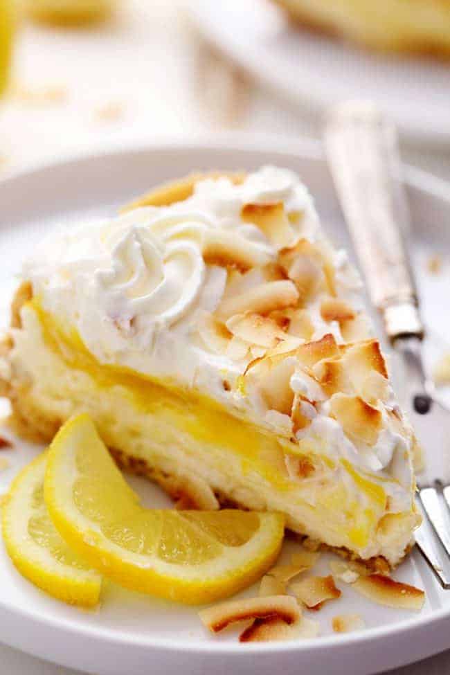 A slice of No Bake Lemon Macaroon Cheesecake on a white plate with a metal spoon and some fresh cut lemon on the side.  