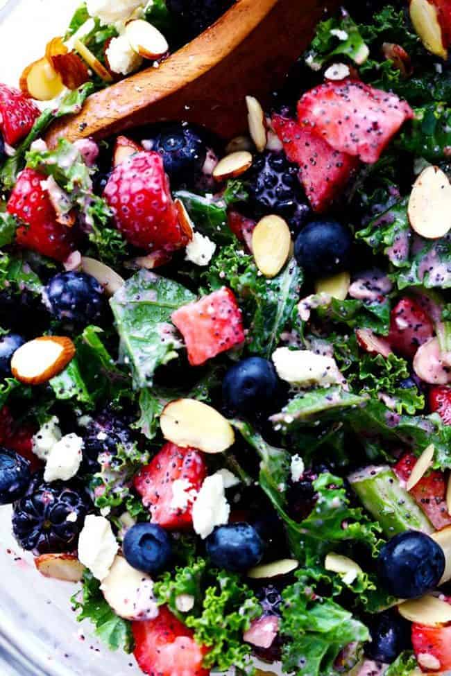 Close up photo of Triple Berry Kale Salad with Creamy Strawberry Poppyseed Dressing in a clear bowl with a wooden spoon. 