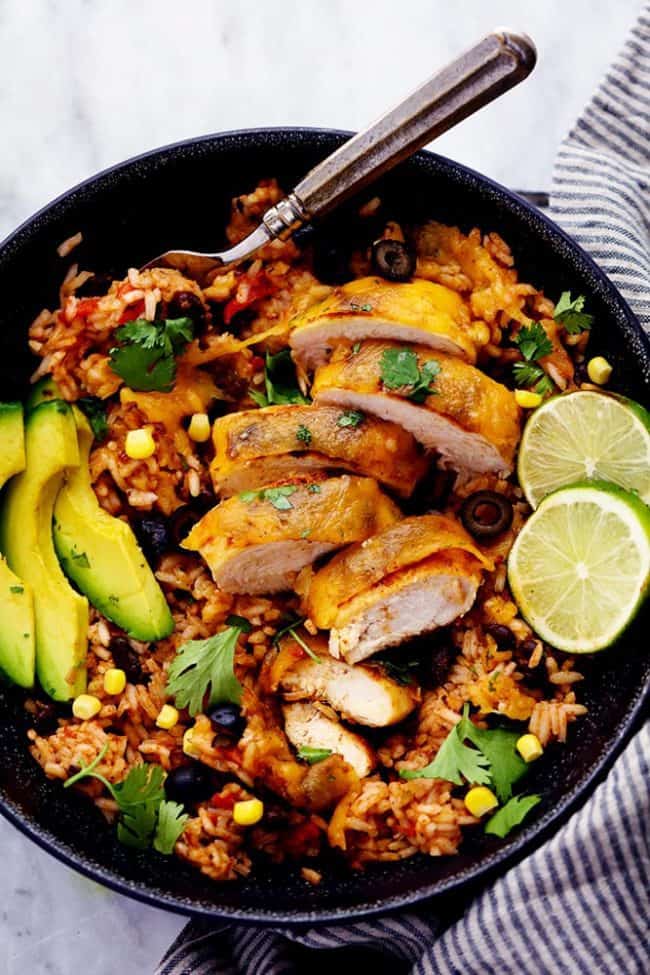 Cheesy Fiesta Chicken and Rice Skillet in a black bowl with slice avocado and limes on top with a metal spoon. 