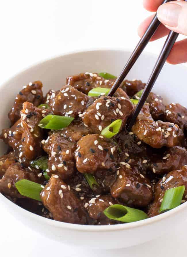 Beef teriyaki in a white bowl garnished with scallions and eating with black chopsticks. 