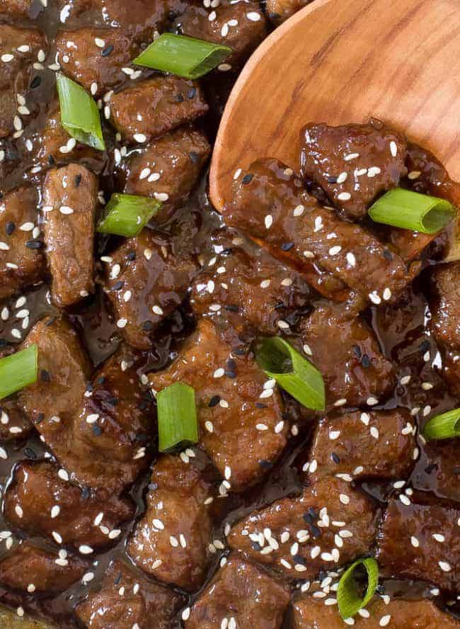 Up close photo of beef teriyaki garnished with sesame seeds and green scallions with a wooden spoon stirring it up.