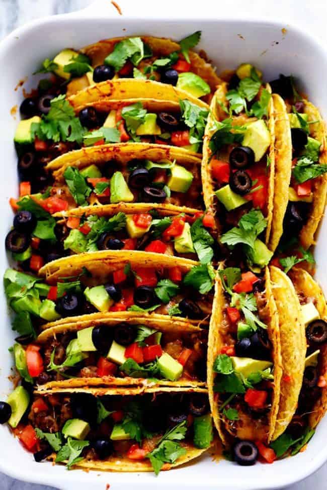 Beefy baked tacos lined up in hard shell tacos garnished with tomatoes, avocados and black olives in a white casserole dish. 