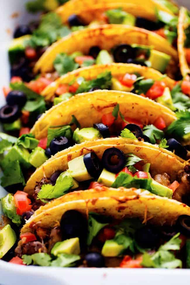 Hardshell tacos lined up in a row garnished with black olives, tomatoes and cilantro on top in a white casserole dish. 