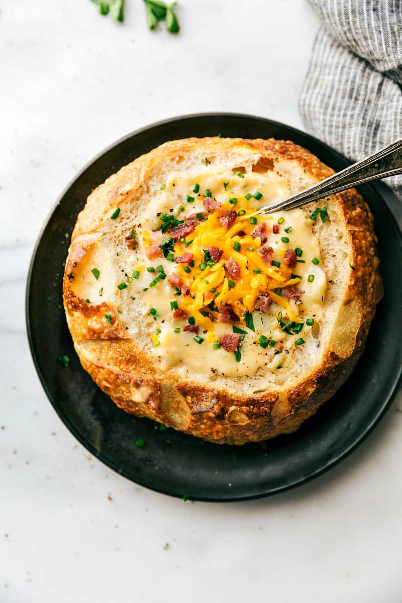 Red Potato Soup in a bread bowl with a spoon in the soup. All sitting on a black plate.