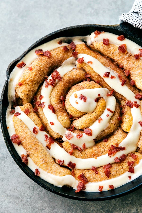 Maple bacon cinnamon roll in a black skillet with frosting overtop and garnished with bacon bits.