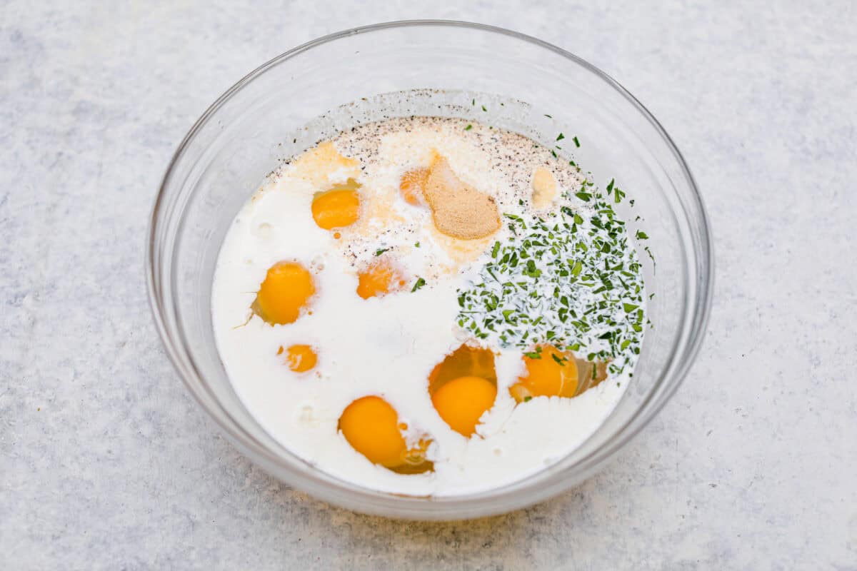 Overhead shot of eggs, half & half, fresh herbs and seasonings in a bowl. 