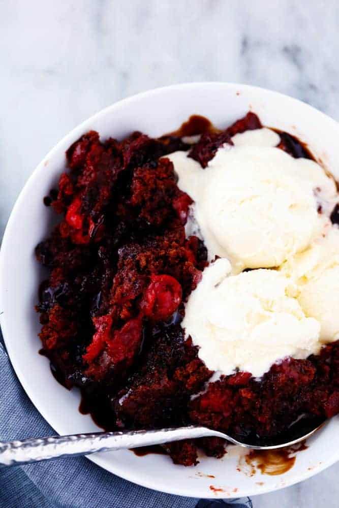 Slow cooker cherry chocolate hot fudge cake in a white bowl with a spoonOn the side with three vanilla ice cream scoops on top.