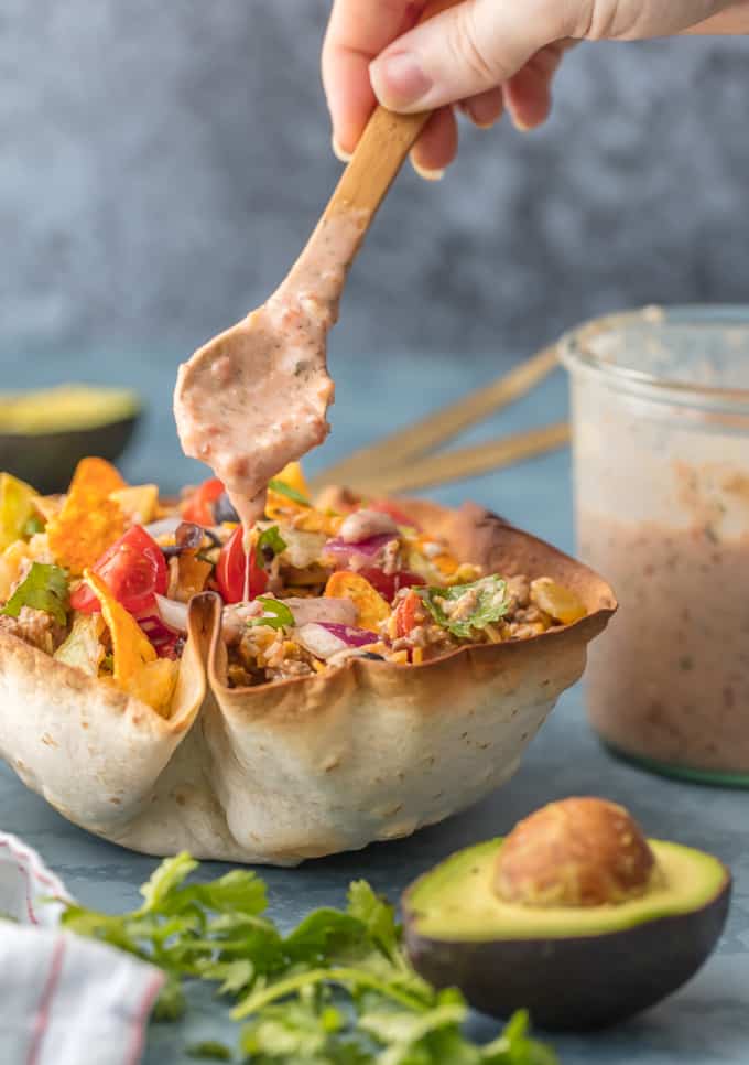 Creamy Taco Salad with refried beans being drizzled over the top. There is a fresh cut avocado in the forefront and a jar of refried beans in the background. 