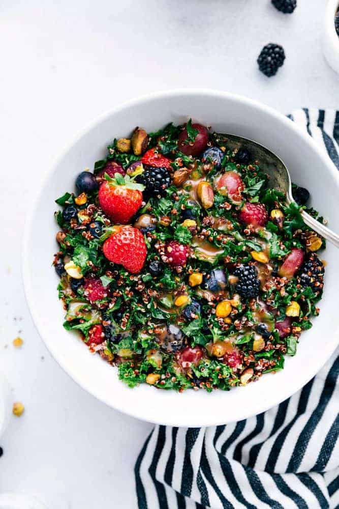 Berry, quinoa, and kale salad in a white bowl with a metal spoon on a black and white striped cloth