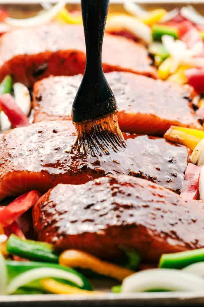 Using a brush to bast uncooked salmon fillets on a sheet pan with sliced green peppers.