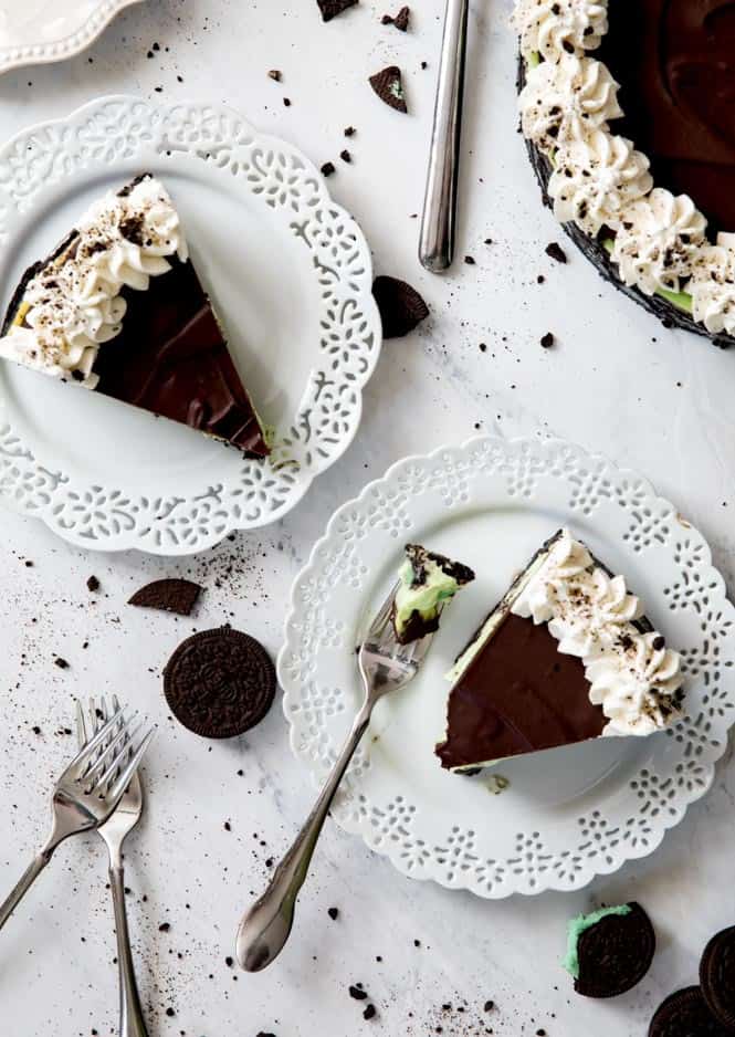 Two slices of Mint Oreo Cheesecake on white plates with metal forks. There are also mint Oreos on the side. 