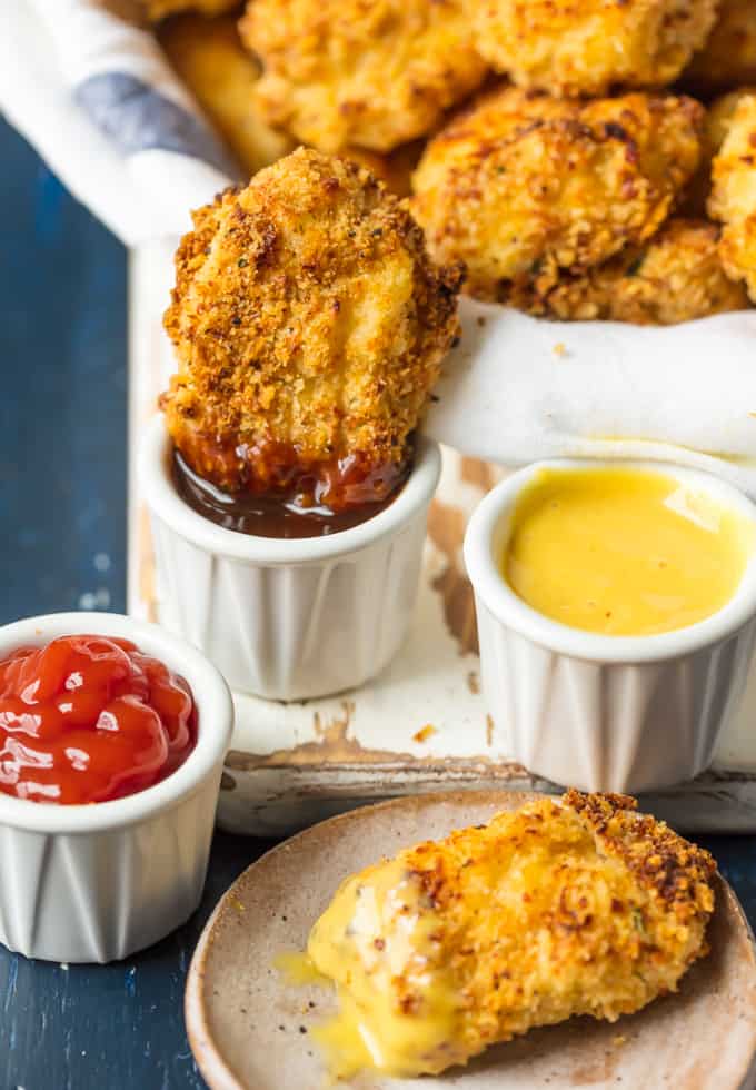 A closeup of Baked Parmesan Chicken Nuggets. One being dipped in BBQ sauce and one being dipped in honey mustard. There is also a small dish of ketchup. 