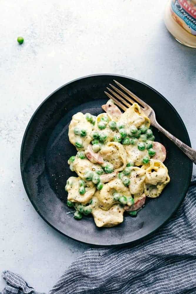 Alfredo Pesto Tortellini with Sausage on a black plate with a metal fork. 