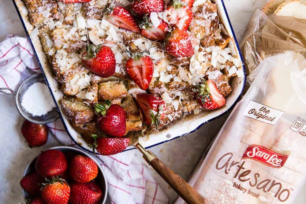 Coconut bread pudding in a white pan with fresh cut strawberries on the side and on top of the pudding. There is a loaf of SarahLee Bread next to the pan. 