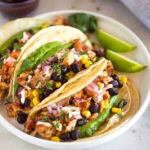 Grilled BBQ Chicken Tacos topped with corn, black beans, avocado, onion and tomatoes, served on a white plate with a small container of extra bbq sauce in the background