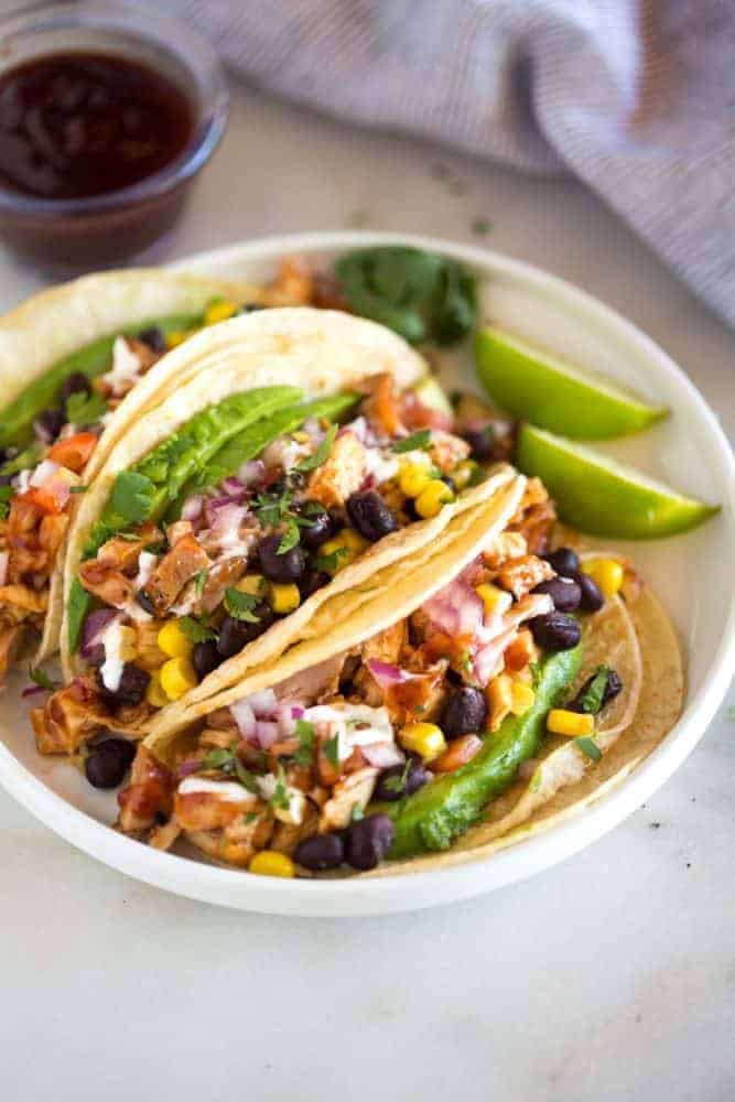 Three Grilled BBQ Chicken Tacos on a white plate with sliced limes on the side. 