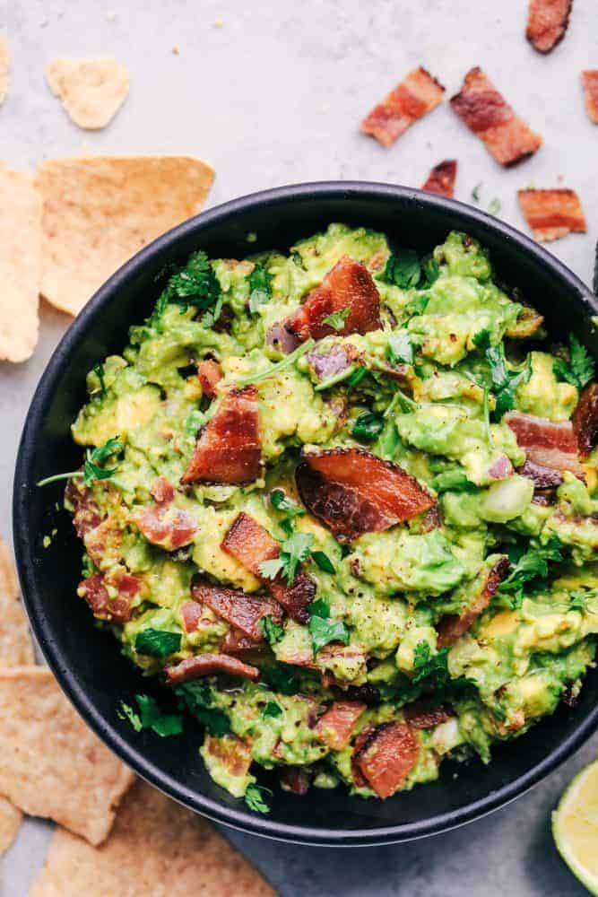 Bacon Guacamole in a black bowl with tortilla chips and bacon bits on the side. 