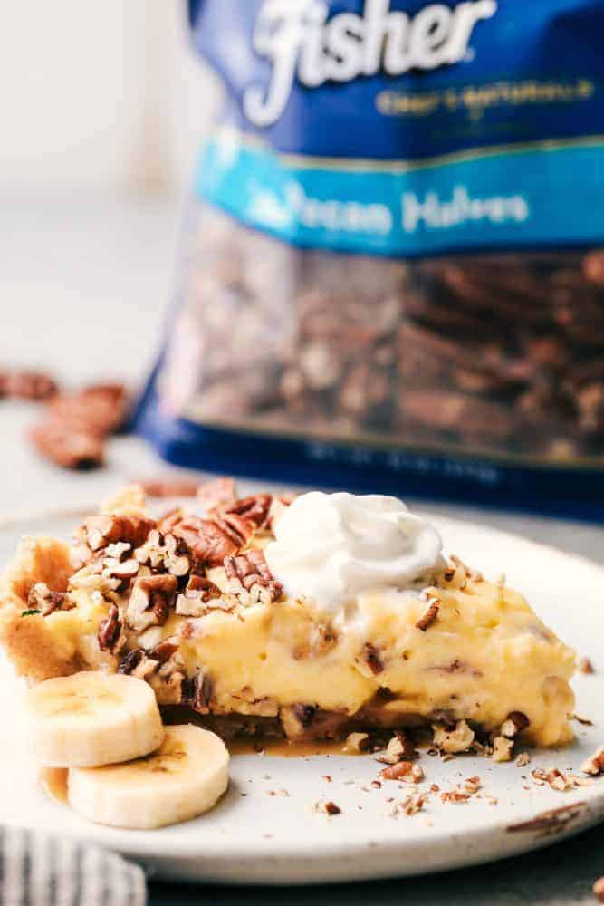 A slice of No Bake Banana Caramel Pecan Pie on a white plate with a couple of slices of fresh cut banana. There is a back of Fisher pecan halves in the background. 