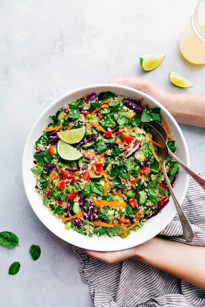 Thai quinoa salad in a white serving bowl with a metal fork and spoon. There are sliced limes on the side. 