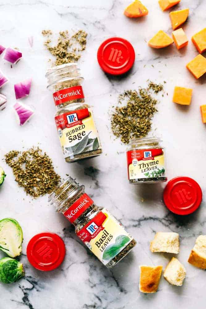 McCormicks seasoning's laid on a white granite countertop spread out with cheese, brussels sprouts and red onion. Rubs sage thyme leaves and basil leaves from McCormick seasoning are spread onto a white granite countertop.